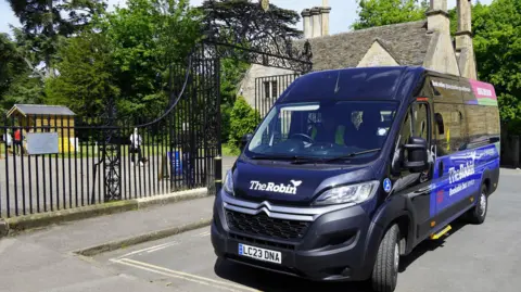 Gloucestershire County Council A  navy mini bus is parked near a black gate with a park in the background and cream-coloured homes.