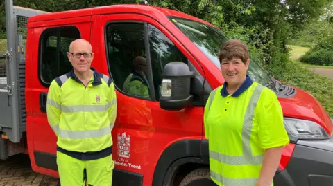 A man and woman in hi-vis tops and trousers stand in front of a red van parked alongside bushes.