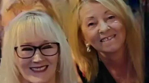 Mandy Moore Two women, one with white blond hair, black glasses and dark lipstick. The other has darker blond hair and gold earrings. Both are smiling at the camera. It is cropped to their faces but looks like they may have an arm around each other.