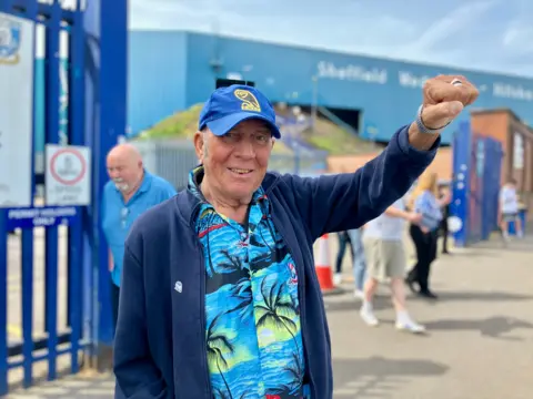 Pedro Marsden standing outside Sheffield Wednesday's ground. He wears a bright blue shirt with palm trees on it, a blue fleeze and a blue Sheffield Wednesday cap. His fist is raised in the air in celebration. 