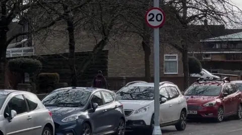 A road of cars in a Stevenage street, showing a 20mph sign, brick houses and a person talking down the street, with a backpack on. 