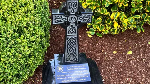 The picture shows a Celtic cross and memorial to Catherine Burns. There are green bushes behind and beside the cross. The memorial says in loving memory of Catherine Burns from Tyrone, Ireland who died at Duffy's Cut in 1832 and was interred in Clonoe in 2015.