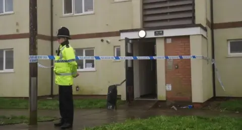 PA Media A police officer with a hi-vis jacket on an a black helmet and uniform trousers, stands guarding the scene at Fownhope Close. Police crime tape is tied to the entrance to a block of flats, and round a telegraph pole, which the officer is standing next to.