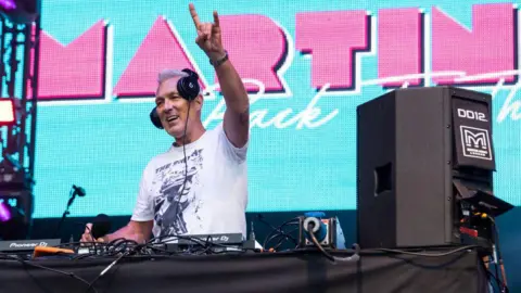 Getty Images Musician Martin Kemp standing behind some DJ decks, wearing headphones and holding a "rock and roll" sign in the air. The screen in the background is blue with MARTIN written in large pink letters.