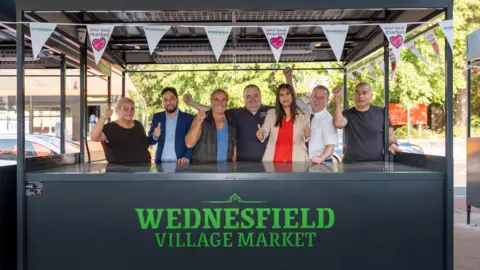 A group of 7 men and women are standing close together, some with their arms around each other, putting their thumbs up. They are looking and smiling at the camera. They are in a black metal market stall with "Wednesfield Village Market" on the front. 
