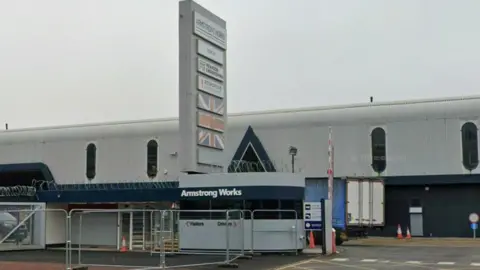 Exterior of the factory, which is large and grey, and the guardhouse which is grey with razor wire around the top. Next to the guardhouse is a tall tower with signs naming the site as Armstrong Works and other company names on it.