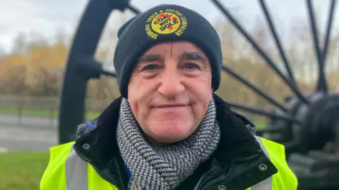 Elizabeth Baines/BBC A man with a black had smiles at the camera. He wears a high-vis and a hat that reads "coal not dole".