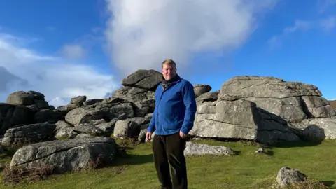 Artist and author, Alex Murdin, is wearing a blue jacket, and stands in front of Bonehill Rocks on Dartmoor. The grass is bright green and the sky is blue with some light clouds. 