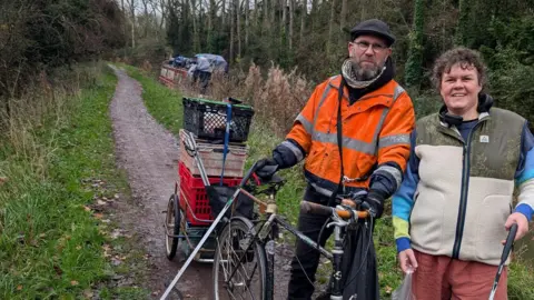 The National Bargee Travellers Association A man in an orange hi-vis jacket and a woman in a winter jacket stand together on a canal footpath near Trowbridge. Both are holding a litter picking tool, while the man is supporting a tricycle contraption with boxes loaded behind the seat.