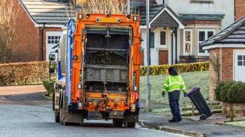 Getty Images A person in a yellow high-vis jacket is seen delivering a black wheelie bin to an orange rubbish lorry