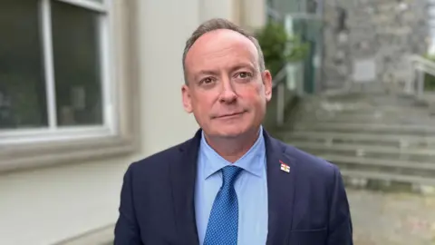 BBC A man with light grey hair wearing a blue suit, a blue shirt and a metallic blue tier looking towards the camera. 