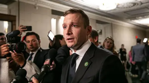 Max Miller, wearing a suit and tie, speaking to reporters holding up microphones in a capitol building