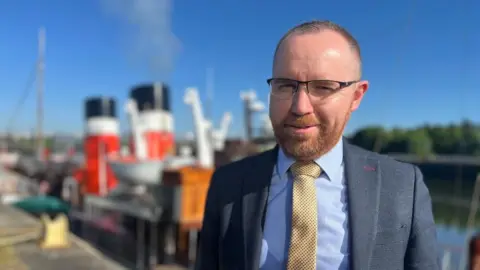 Paul Semple, General Manager of Waverley Excursions, smiles at the camera while standing in front of the paddle steamer. He wears a navy suit with a blue shirt and yellow tie. 