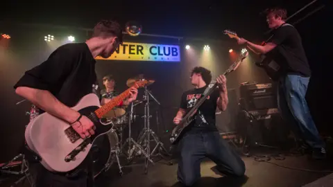 Seymour Quigley performing with his band on stage at the Hunter Club. He is crouching down while holding his bass guitar and looking back towards his drummer. There are two other guitarists either side of him, both of whom are looking back towards the drummer.