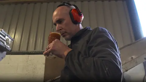 Ty Gwyn Cider Culpin, wearing a grey fleece and orange headphones, brings a glass of cider up to his nose to smell.