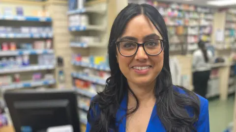 Jasvinder Kaur Lali a Superintendent Pharmacist standing at the chemist shop counter of the Carlton Hill Pharmacy in Nottinghamshire. She is wearing a blue jacket and white blouse 