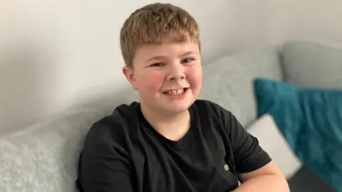 A smiling boy is sat on a sofa, looking at the camera with his arms folded across his chest. He is wearing a black t-shirt and has short medium brown hair with a fringe. 