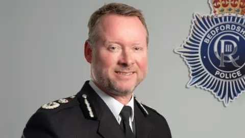 Bedfordshire Police Chief Constable Trevor Rodenhurst is wearing a police uniform and smiling. Behind him on the white wall is a badge for Bedfordshire Police