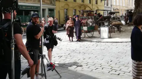 BANES A market is taking place on a cobbled street, with people milling around, while a film crew, with people dressed in modern clothing, film. 
