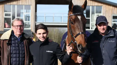 Darren Staples / AFP via Getty Images Race horse owner Harry Redknapp, trainer Ben Pauling and Jockey Ben Jones pose with Cheltenham Festival hopeful The Jukebox Man at the Ben Pauling Stables. There is a wood-panelled building behind them which is split in two and connected by a glass balcony walkway. It is a sunny day.