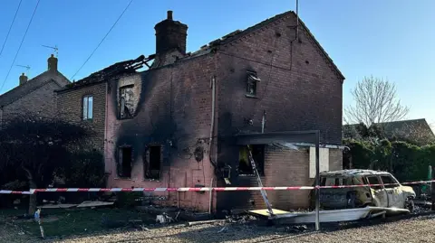 BBC/Joe Weir A side angle of two semi-detached brick houses badly affected by a fire. The one closest to the camera has pink bricks with the windows burnt out, and the roof is missing. There is a burnt-out car on the right and a tree in front with a red and white cordon tape wrapped around it, stretching across the front lawn and the car. The neighbouring house has brown bricks and its roof is partially missing