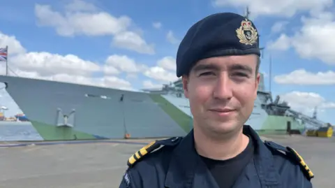 Lieutenant commander Dominic Jacob has his Royal Navy uniform on. The large grey ship with green detailing is docked in the background.