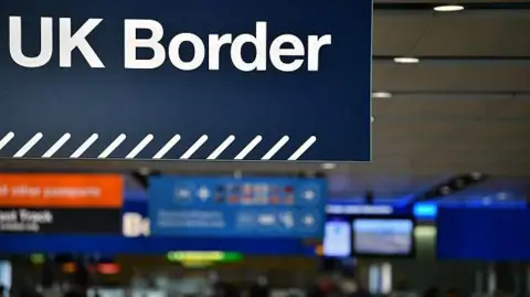 Getty Images A close-up of a blue sign with white text that reads "UK Border" hanging from a ceiling. Below the text is a diagonal striped border in white. The background shows a blurred airport terminal with various smaller directional signs in orange and blue.
