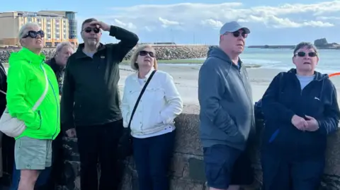 People look up at the sky watching the Jersey air show. There are five people leaning against a wall. One man has his hand over his eyes to shade his face from the sun.
