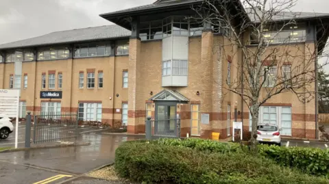 A brown brick three storey building with a sign that says SpaMedica. There are car parking spaces in front of the building and a hedge and tree.