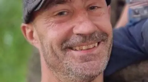 Stefan Watkins is smiling at the camera wearing a grey cap, with grey stubble, blue eyes and dark eyebrows. He has laughter lines around his eyes and is tilting his head.