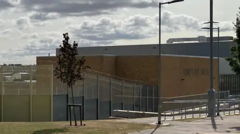 An outside view of HMP Five Wells prison. It shows a large brick building surrounded by metal fencing. 