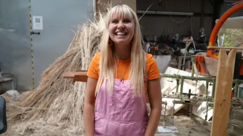 Shaun Whitmore/BBC A woman stands inside a building where long crops that have been cut from a field rest behind her. She is smiling and has long blonde hair. She is wearing an orange T-shirt with pink dungarees over the top. 