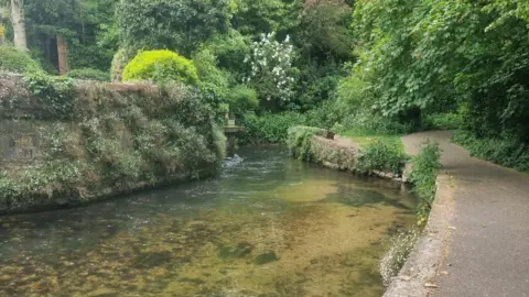 Ian Griffiths View from the footpath alongside the River Frome in Dorchester. The clear, shallow river runs between two stone walls with a path on one side and is surrounded by trees and greenery.