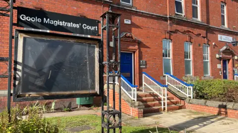 The exterior of Goole Magistrates’ Court - a red-brick building with a blue entrance door and steps with blue handrails. In the foreground, a black-framed sign reads “Goole Magistrates’ Court,” with an empty noticeboard, set beside a small lawn and paved walkway.