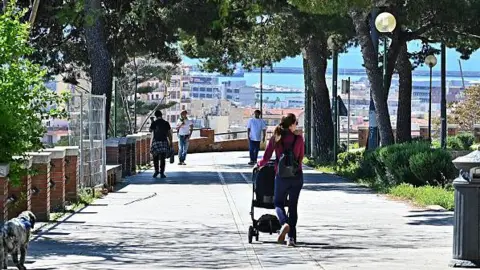 A woman pushes a pram as people walk along Viale Enrico Endrich in Cagliari, Sardinia, Italy