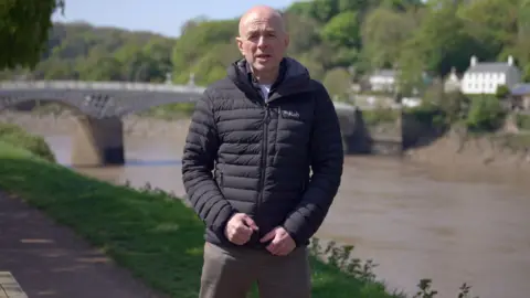 Gareth Lewis standing on a riverbank with a bridge in the background