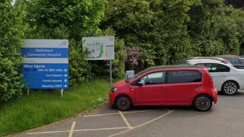 A line of cars parked outside Haselmere Community Hospital,  parked beside the hospital's sign.