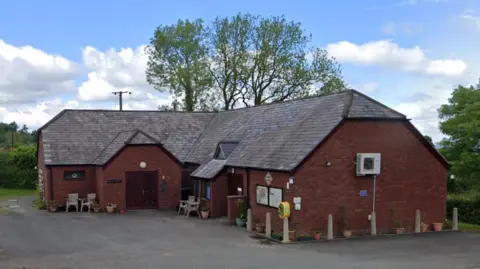 Google An L-shaped red brick building with a large dark roof, surrounded by bollards and a car park.