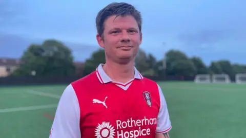 Simon Thake A man with short dark hair stands on an empty football pitch wearing a red strip