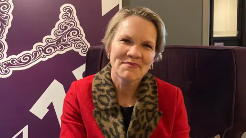 A close up of Sasha Kazantseva-Miller. She has short fair hair, gold earrings and is wearing a red jacket.