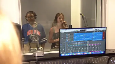 Two female students are standing behind a panel of glass in a recording booth. The one standing to teh left wears an NFL football t shirt which is a dark blue with the writing New York in red and the logo of a football helmet on, she has black braided hair tied back in an up-do and black headphones on, with black thick framed glasses and she is standing behind a microphone. The other performer stands to the right, she has long curly brown hair and a pait of black headphones on. She is holding her microphone and wears a strapless black and sequinned top. In the foreground of the picture is a computer screen which shows music editing software.