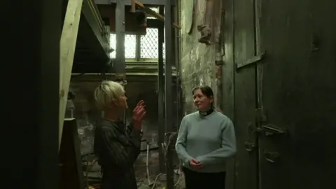 Two women standing in conversation in a gloomy part of a church between a large door and the remains of the wooden structure of a pipe organ, which is being dismantled. The woman on the right has short blonde hair and wears a dark shirt. The woman on the right has brown hair, tied back, and is wearing a light-blue jumper over a vicar's black shirt and white dog-collar. A small stone gargoyle can be seen on the wall above her head. Light shines through a window.