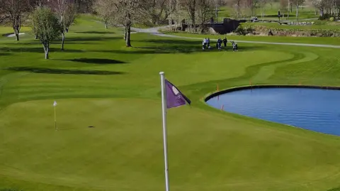 Official Hole in 1 A golf ball trickling into the hole on a golf course. Water can be seen to the right and a purple flag with the Carus Green Golf Club name on it.