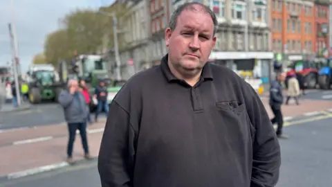 Christopher Duffy is standing in a street in Dublin. He's wearing a black long-sleeved polo shirt. People are in the background but are out of focus. 