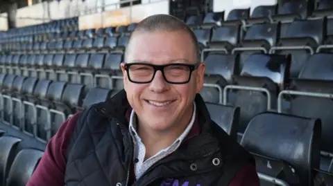 Tom Williams/BBC A man with glasses, wearing a maroon top, white shirt and a black gilet, sitting in a football stadium stand and smiling