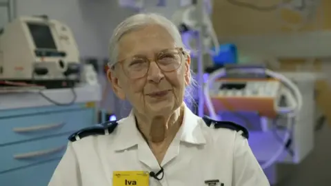 Nurse Iva, wearing a white uniform with a yellow badge that says Iva in black, bold text, smiles to camera.