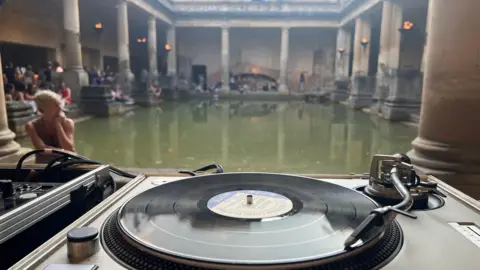 A deck with a vinyl playing is in the foreground. In the background is a Roman thermal bath, which looks like an open-top green rectangular pool, with stone pillars surrounding it. There are people sitting around the pool. 
