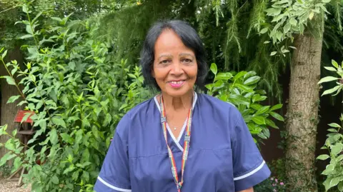 A woman in purple scrubs smiles at the camera. She wears her black hair up and has a multi-coloured striped NHS lanyard around her neck as she stands in a garden