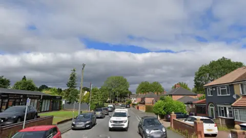 Google Streetview image of Springhill Lane. Cars are parked either side of the road with a white pick-up  coming towards the camera in the centre. There are businesses on the left and houses on the right