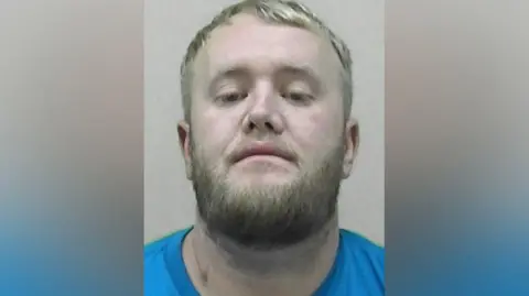 Northumbria Police Mugshot of McMahon. He has a thick fair beard and short blond hair and is wearing a blue t-shirt.